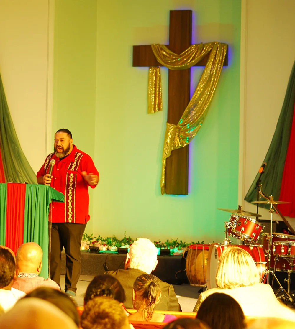 Pastor preaching to the congregation at Grace and Praise Bangladeshi Church with the cross behind him.